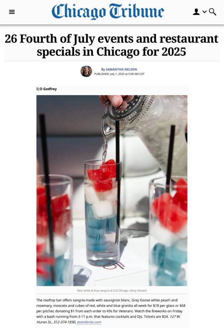 A close-up of a red, white, and blue sangria being poured at I|O Godfrey rooftop bar in Chicago. The drink features layers of colored granita cubes in a tall glass with a black straw, as clear liquid is being strained from a cocktail shaker. Other glasses of the patriotic-themed sangria are visible in the background.